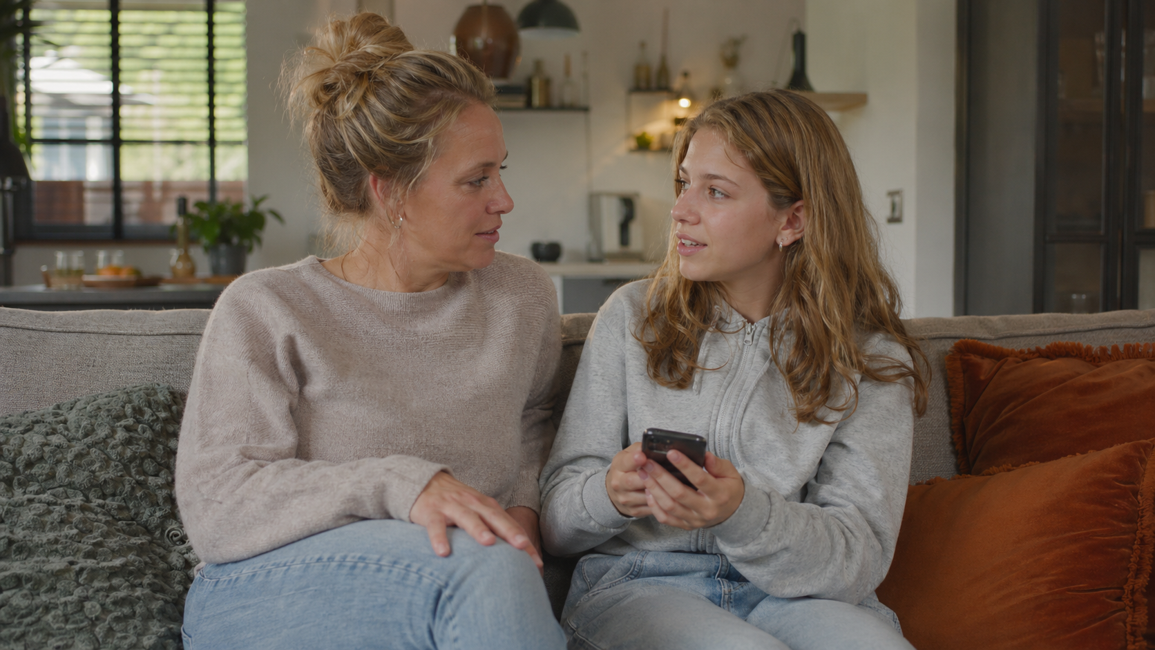 Moeder en tienerdochter zitten samen op de bank en praten over de smartphone in haar hand.